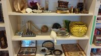 Wide view of three shelves showcasing a variety of decorative items including framed art, shells, vases, and brassware.