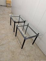Two black metal frame side tables with beveled glass tops, shown on a concrete floor against a white wall.