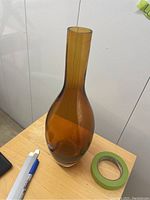 Amber glass vase with heavy bottom sitting on a wooden surface alongside a roll of green painter's tape and some markers.