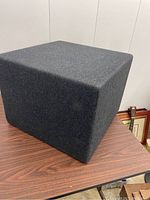 Front and side angle of dark gray wool fabric cube ottoman on wooden table, showing the smooth texture and clean edges with slightly rounded corners.