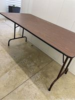 Table side view showing wood grain top and folding metal legs on labeled textured floor.