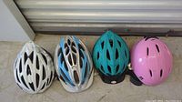 Four helmets aligned in a row on ground; includes white, blue-white, teal-black, and pink helmets.