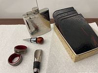 Overall view of stainless steel flask, bottle stoppers, and napkin rings on white cloth, coaster box to right.