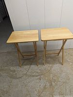 View of two foldable wooden tables standing side by side on a concrete floor in front of a white paneled wall.