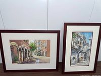 Two framed watercolor paintings depicting European streets with cafes and laneways. One depicts a street with arches and outdoor seating, the other shows a narrow laneway with balconies and arch.