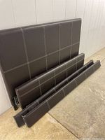 Photo showing brown upholstered headboard, footboard, and two side rails laid against the wall on a concrete floor.