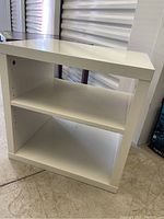 White rectangular Ikea Corras side table with one adjustable shelf and smooth surface, standing on a concrete floor.