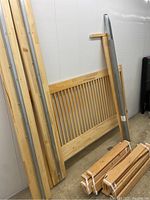 Photo showing disassembled wooden bed frame parts leaning against a white wall, including slatted headboard, side rails with metal reinforcements, and tied bundles of wooden slats.