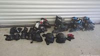 Four pairs of inline skates with assorted protective gear including gloves, knee pads, and arm/elbow pads arranged in a row against a corrugated metal backdrop.