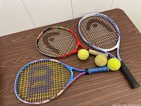 Three kids tennis rackets (blue, red, purple) and three tennis balls arranged on a table.