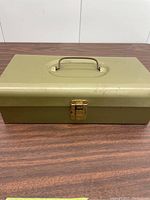 Closed view of vintage green metal propane case with brass latch and handle on top, placed on wood surface.