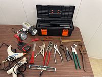 Full lot showing toolbox, Milwaukee flashlight, glue guns, small hammer, pliers, wrenches, clamp, and spirit level on wooden table.