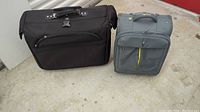 Photo of both luggage pieces side by side on floor; one black larger wardrobe suitcase and one smaller grey-yellow carry-on suitcase.