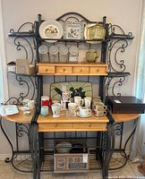 Full view of bakers rack loaded with mugs, dishes, glass jars and other kitchen items, showing four small drawers, two larger drawers, wooden side shelves, and metal frame with scroll details