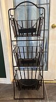 Front view of the Mesa three-tier wrought iron basket stand showing all three baskets and the stand's frame against a windowed door.