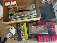 Box with assorted BBQ tools including spatulas, tongs, grill baskets, starter fluid bottles and wool blanket.
