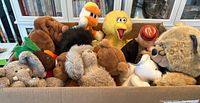Box of assorted plush toys including Sesame Street characters Big Bird and Elmo, various dog plushes, a moose, duck, polar bear, and a toy figure with a red dome head.