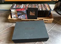 Photo showing a box containing multiple DVD cases including The Godfather DVD Collection in a black slipcase, two PlayStation 2 game cases - NASCAR 06 and Gran Turismo 3, and various other DVDs lined up