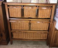 Front view of wood cabinet with three smaller wicker baskets on top row, two medium baskets in middle, one large basket bottom drawer. Leather style handle pulls on all baskets.