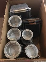 Box containing ten stainless steel canisters with metal locking clasps and clear plastic lids, stacked with two wood baskets and two metal mesh baskets visible at top of box.