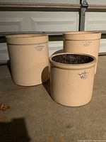 Three beige stoneware crocks stamped with a blue crowned logo and size numbers, displayed upright on concrete floor.
