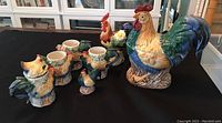 Photo showing six items including Fitz and Floyd rooster lidded bowl, four mugs with rooster designs, and unmarked rooster figurine on black surface