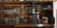 Wooden cabinet shelf containing multiple vintage glassware items including wine glasses, clear drinking glasses, ashtrays, a boxed wine bottle accessory set, and a collectible race car lighter.