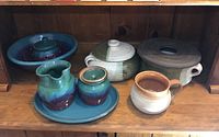 Display of multiple glazed pottery pieces including blue plate with small pitcher, lidded bowls and pottery mug.
