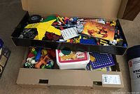 Wide shot of open box showing assorted Lego pieces, rubber animal figures, party tablecloths in packaging, kids party game kits, and a white container with plastic lid.