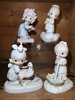 Four Precious Moments porcelain figurines displayed on wood shelf, including one with umbrella and ducks, an angel figurine, and children with animals and toys.