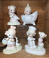 Five Precious Moments figurines displayed in wood shelf including a girl holding a goose, child in bathtub, child with nurse cap and a child with teddy bear.