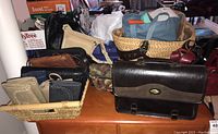 Overview of fifteen handbags, purses, and wallets laid on table including woven basket bag, leather wallets, and satchel.