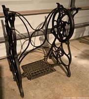 Side view of black cast metal sewing machine stand showing treadle pedal and pulley wheel with decorative scrollwork.