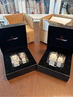 Two black Swiss Time branded watch boxes each containing one men's and one women's watch in silver-tone stainless steel with rectangular faces, displayed with original outer cardboard boxes in the background.