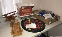 Wooden toy truck, wooden airplane, dartboard with safety darts, crayons, table tennis paddle, pencils, and Jammin Drum arranged on two tables in natural light.