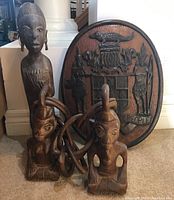 Photo of three wood carvings from Jamaica - one oval coat of arms wall hanging and two human figurines all carved from wood.