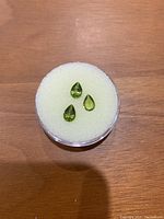Three pear cut green peridot gemstones displayed on a white foam pad inside a circular plastic case, showing size and color.