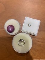Three loose gemstones on foam pads: large round cabochon purple/red stone, small round faceted yellowish stone in square box, large square cushion-cut faceted pale stone in round container.