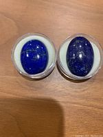 Two oval lapis lazuli stones with polished surfaces and gold flecks, displayed side by side in round cases on wooden surface.
