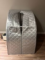 Silver quilted portable sauna enclosure with front zipper and pocket visible, pump is seen in background.
