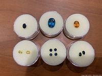 Six sapphire gems displayed in round clear containers on foam: one dark blue oval, one bright blue oval, one golden yellow, two light yellow ovals, four small blue rounds, two blue pears