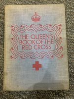 Front cover of 'The Queen's Book of the Red Cross' showing beige fabric cover with red title and Red Cross emblem