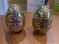 Front view of two small gold-tone decorative eggs with raised colorful floral and leaf patterns in enamel, shown standing upright side-by-side on a wooden surface.