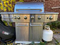 Front view of the Amana barbecue grill showing four main burner control knobs, two doors below, and one propane tank to the right side.