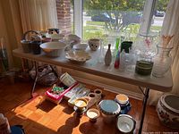White resin folding catering table with various glassware and ceramics placed on top and underneath for staging purposes, visible parquet wood floor and sunlight from window.