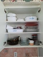 Kitchen cabinet shelf with stacked Corning Ware casserole dishes and Pyrex glassware with lids, along with several ceramic ramekins.