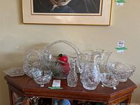 Full view of eight clear crystal and glass pieces on a wooden table, includes a handled basket bowl with faux fruit, pitcher, bowls, and vase.