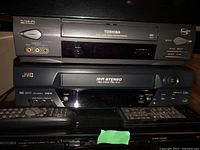 Toshiba Hi-Fi Stereo VCR stacked atop JVC Hi-Fi Stereo VHS player with two remotes on glass shelf