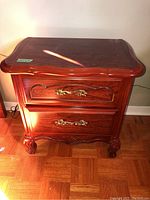 Front view of first wooden end table or night stand with two drawers showing gold-tone drawer handles and carved legs.