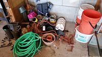 Wide shot of the lot showing green garden hoses, plastic buckets, garden art, bird feeders, and extension cord on concrete floor in garage corner.
